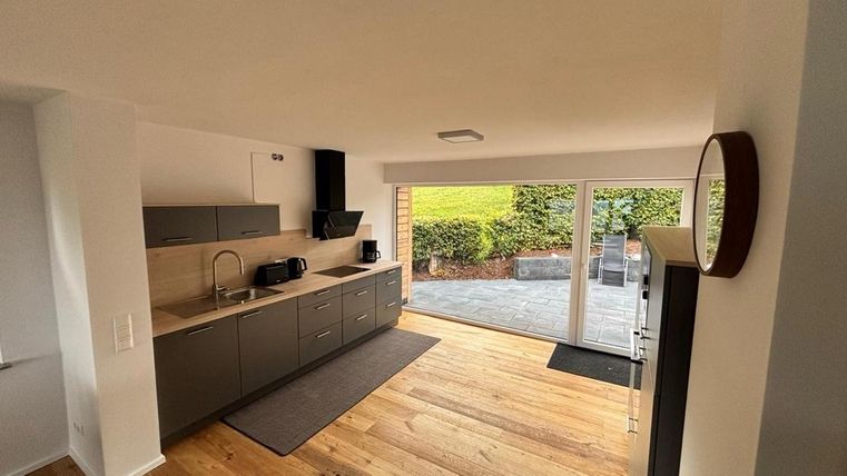 A modern kitchen with gray cabinets and wooden flooring. Large windows provide a view of the outdoor area with a garden.