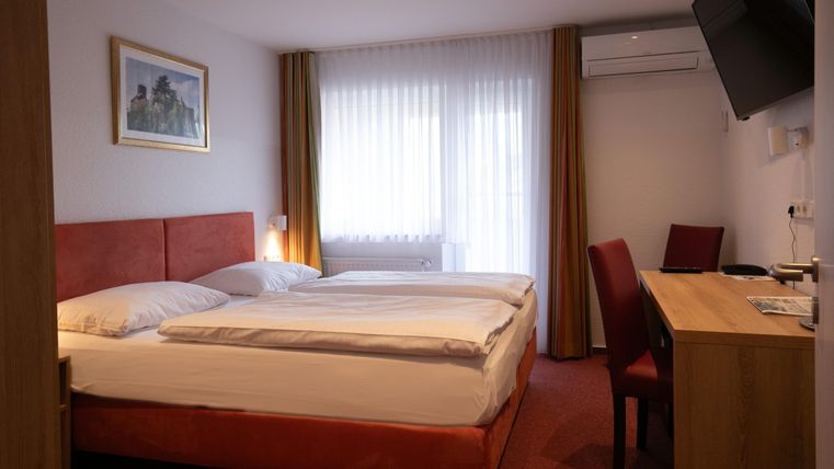 Ein gemütliches Hotelzimmer mit einem Doppelbett und einem Schreibtisch. Große Fenster sorgen für viel Tageslicht.