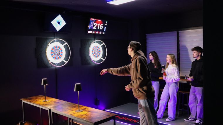 A person is playing darts in a modern room with digital dartboards. In the background, three other people are standing and watching the game.