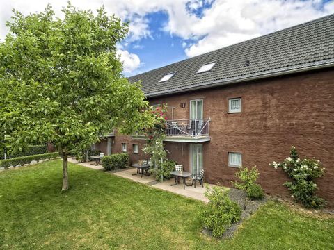 Ein gemütliches Wohngebäude mit roter Fassade, umgeben von einem gepflegten Garten. Im Vordergrund stehen Tische und Stühle unter einem Baum.