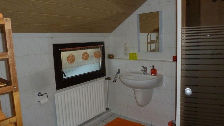 A small bathroom with a sink, a window, and a shower.
The floor is equipped with white tiles and an orange carpet.