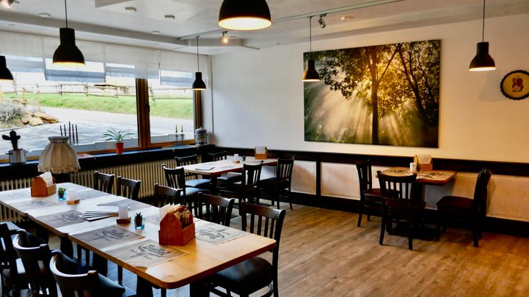 A cozy restaurant with wooden tables and chairs. A large picture hangs on the wall depicting sunlight streaming through trees.