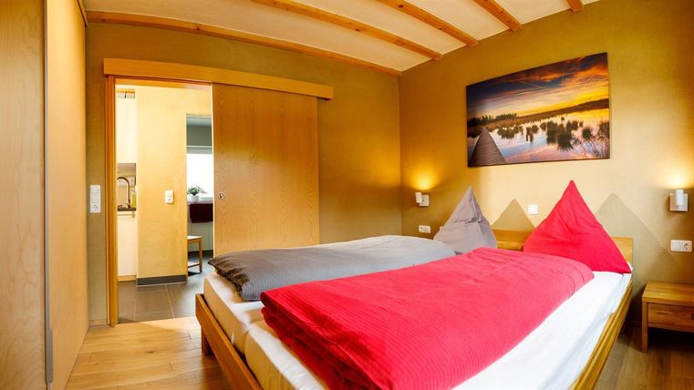A cozy bedroom with a double bed and red pillows. The walls are painted in a warm yellow tone, and a window provides daylight.
