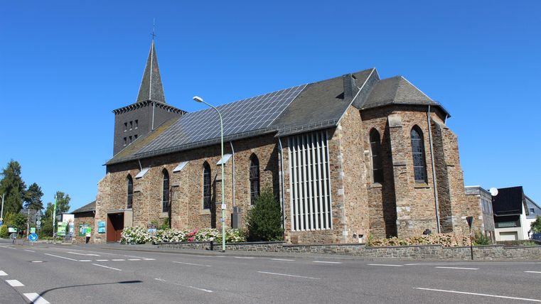 Eine alte Steinkirche mit einem spitzen Turm steht an einer Straße. Der Himmel ist klar und blau.