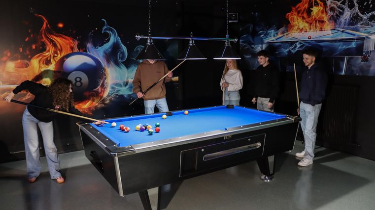 A group of four people is playing billiards on a blue table. Colorful, dynamic graphics can be seen in the background.