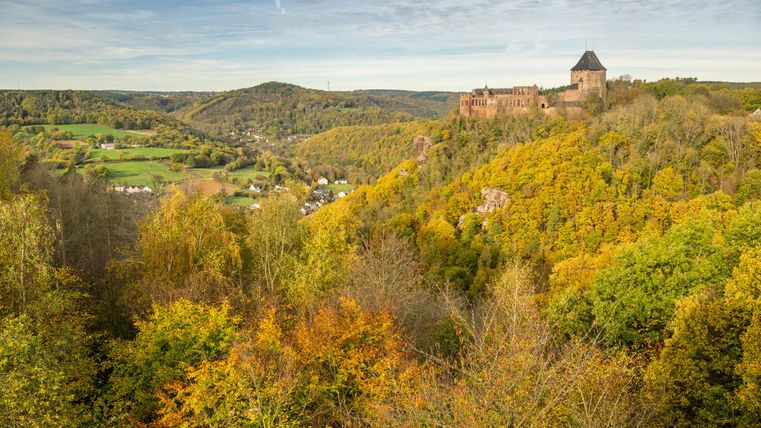 An impressive castle ruin on a hill, surrounded by colorful autumn trees. In the background, gentle hills and a green landscape extend.