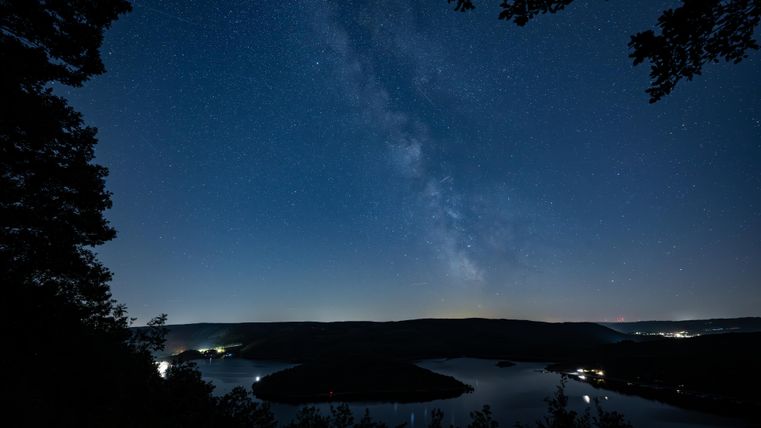 Ein nächtlicher Himmel voller Sterne mit der Milchstraße. Im Vordergrund ist ein ruhiger Fluss und sanfte Hügel zu sehen.