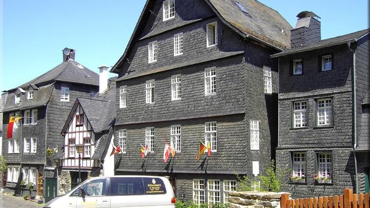 Ein historisches Gebäude mit schwarzer Holzverschalung und vielen Fenstern. Im Vordergrund steht ein Van und es sind mehrere Fahnen zu sehen.