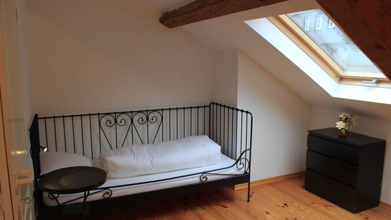 Een gezellige kamer met een ijzeren bed, een kleine tafel en een ladekast. Het licht valt door een raam in het schuine dak.