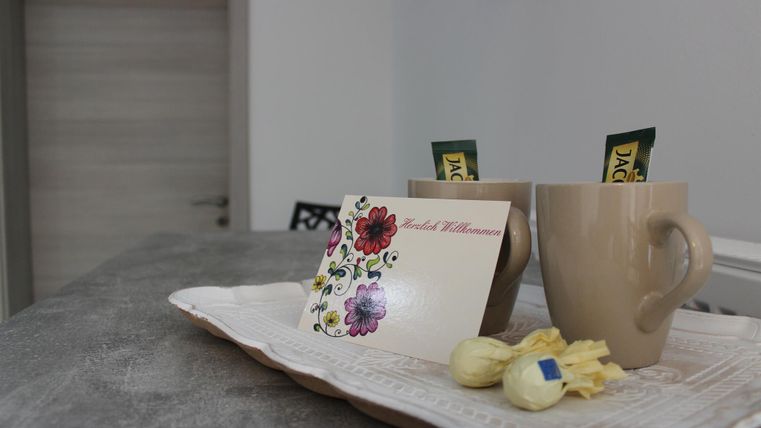 Two cups on a tray, accompanied by a welcome card and small sweets. A bright room can be seen in the background.