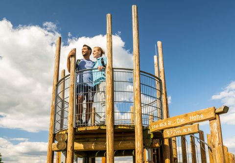 Ein Pärchen steht auf einem Aussichtsturm und schaut in die Ferne. Der Himmel ist blau mit einigen Wolken.