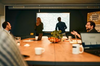 Ein Team in einem Besprechungsraum brainstormt an einer Wandtafel. Auf dem Tisch stehen ein Obstkorb und Kaffeetassen.