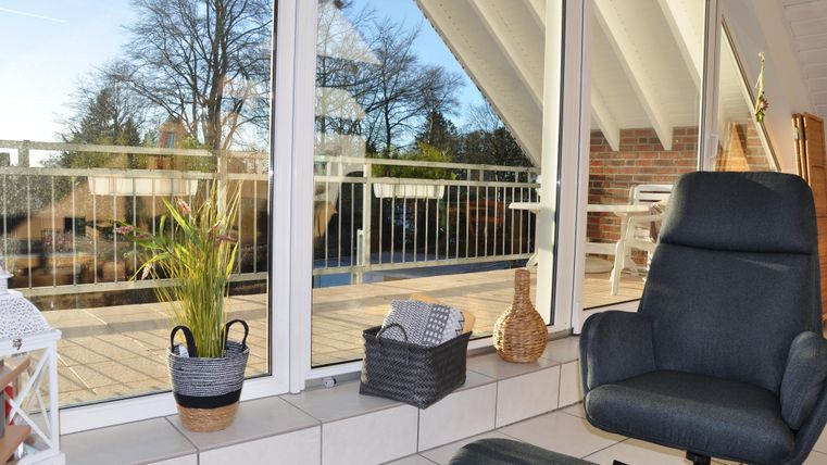 A cozy room with large windows that lead to a balcony with an outdoor view. In the foreground, there is a comfortable armchair and some plants in baskets.