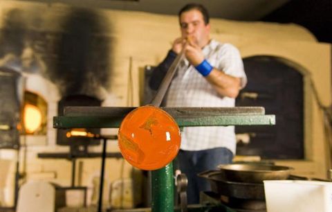 Een glasblazer werkt aan een glasstuk in een atelier. Op de achtergrond zijn ovens en gereedschappen te zien.