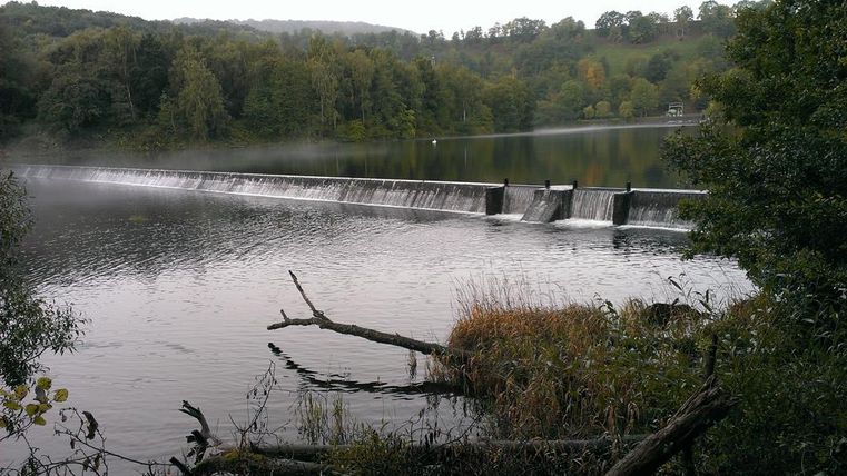 A dam stretches across a tranquil lake, surrounded by trees and gentle hills. The waterfall flows gently over the edge of the dam.