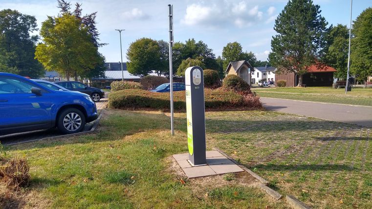 Een laadstation voor elektrische auto's staat op een parkeerplaats. Op de achtergrond zijn bomen en een rustige woonwijk te zien.