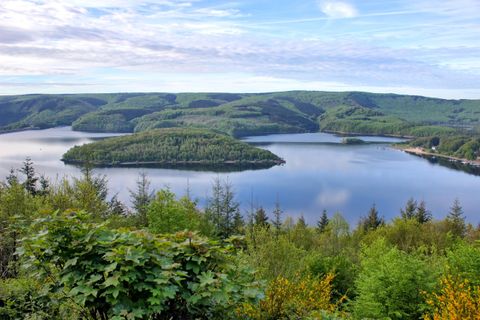 A picturesque lake surrounded by green hills and trees. The sky is partly cloudy and reflects a serene atmosphere.