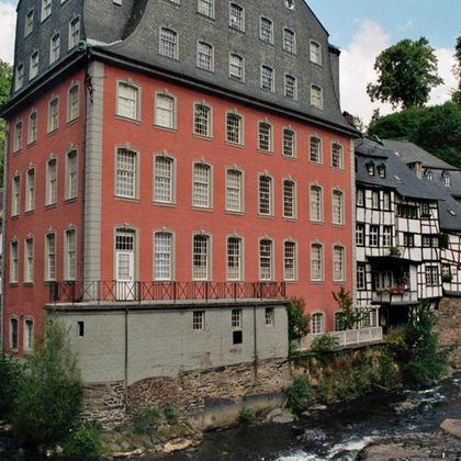 A red building next to a river in an idyllic setting.
Surrounded by traditional half-timbered houses and green trees.