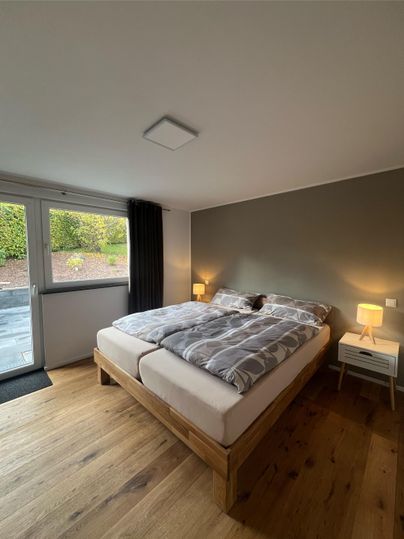 A cozy bedroom with a large bed and a wooden floor. There are windows looking outside and two nightstands with lamps.