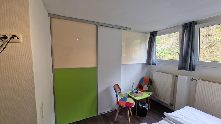 A modern room with a green wardrobe, a small table, and colorful chairs. The windows offer a view of nature.