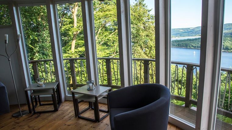 A cozy room with large window fronts that offer a view of a forest and a lake. There are comfortable chairs and tables that create a relaxed atmosphere.