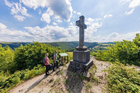 Eine Gruppe von Personen steht an einem Kreuz auf einem Hügel mit Blick auf die grüne Landschaft. Der Himmel ist klar und mit einigen Wolken versehen.
