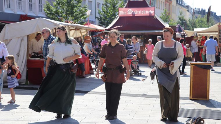 Drei Frauen in historischer Kleidung gehen über einen Marktplatz. Im Hintergrund sind Marktstände und viele Menschen zu sehen.