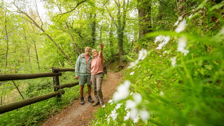 Ein Paar steht lächelnd auf einem Wanderweg in einem grünen Wald. Um sie herum blühen weiße Blumen und die Atmosphäre ist frisch und einladend.