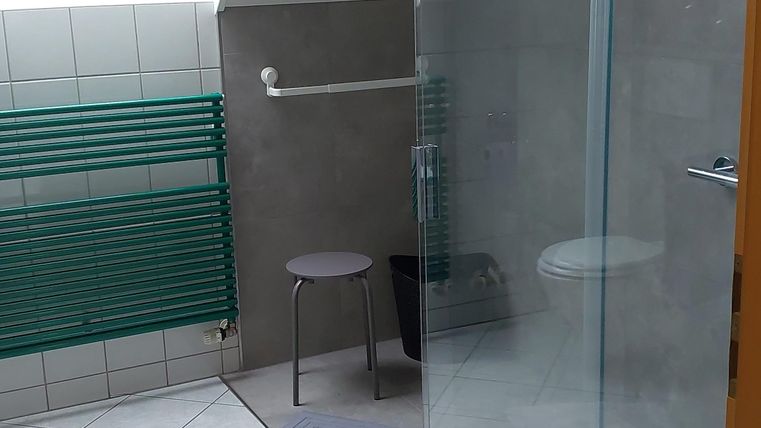 A modern bathroom with a glass shower and a small stool. The space has a bright, friendly atmosphere.