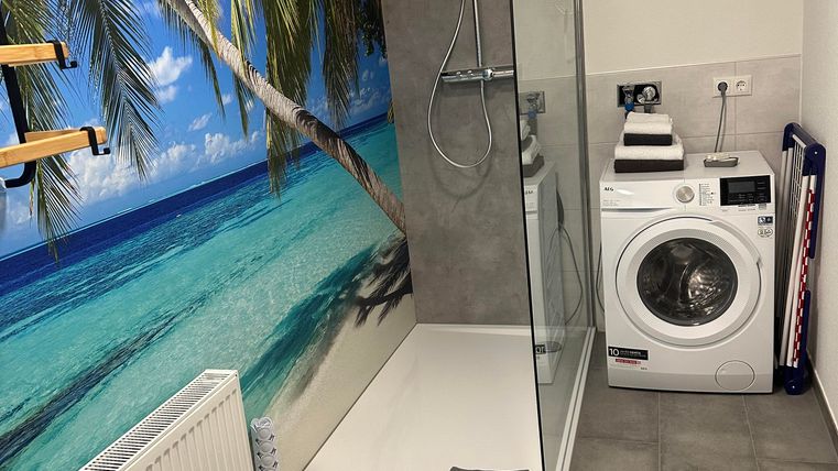 A modern bathroom with a walk-in shower and a washing machine. On the wall, there is a tropical beach motif with palm trees.