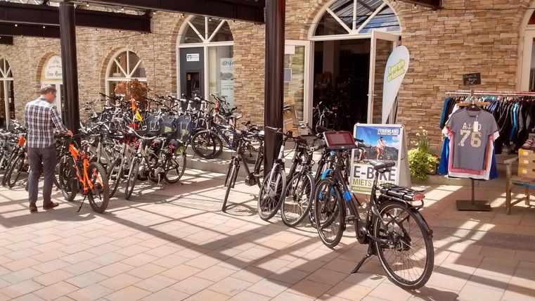 Een grote selectie fietsen staat in een groen binnenhof. Op de achtergrond zijn mensen en winkels zichtbaar.