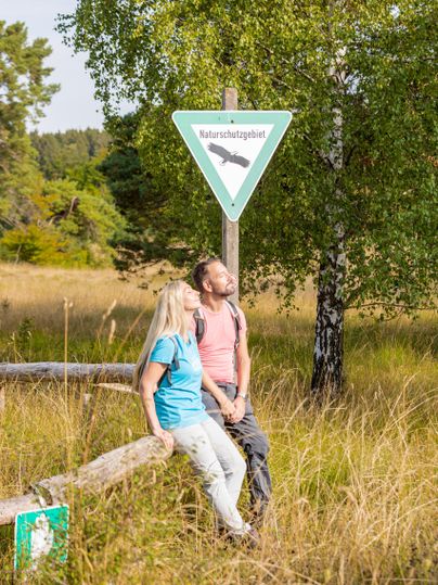 Ein Paar sitzt entspannt auf einem Baumstamm in der Natur. Im Hintergrund steht ein Schild, das ein Naturschutzgebiet anzeigt.