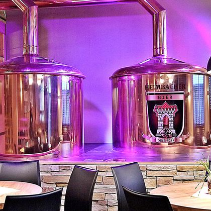 Two large copper kettles for the brewery are standing in front of a purple illuminated wall. The kettle on the left side features the logo of a brewery.