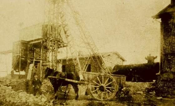 Een oud beeld van een bouwplaats met een stoomlocomotief en een puttenbouw. Een paard trekt een wagenlading in de buurt van de werkplek.