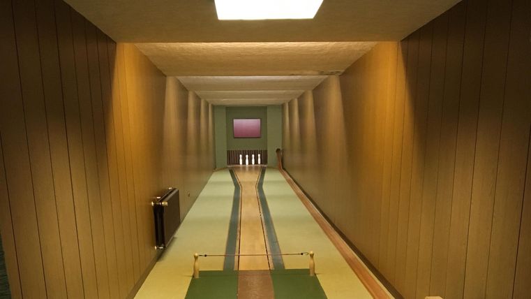 An elegant bowling alley with wooden paneling and soft colors. At the end of the lane, pins and a light area are visible.