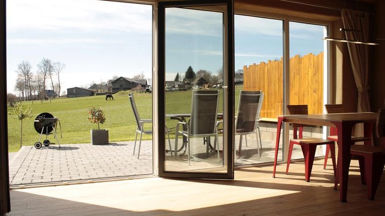 An inviting interior overlooking a green meadow and houses in the background. Outside, chairs and a grill are ready.