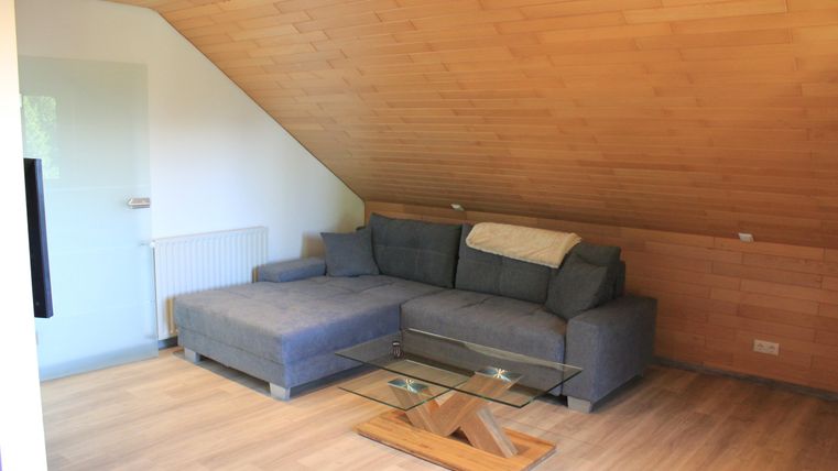 A cozy living room with a gray couch and a glass table. The walls are paneled with wood, and the floor is made of light wood.