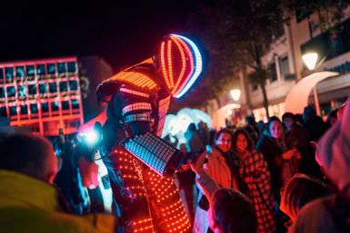 Ein farbenfroher Tanzender in einem leuchtenden Kostüm steht im Mittelpunkt einer Menschenmenge bei Nacht. Die Atmosphäre ist festlich und lebhaft.