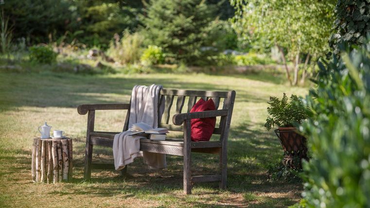 Een gezellige bank in de tuin met een rood kussen en een deken. Naast de bank staat een kleine houten tafel.