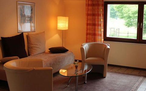 A cozy living room with a sofa, two armchairs, and a glass table. The room has large windows and pleasant light.