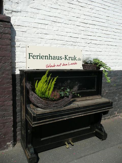Een oud piano versierd met planten staat voor een witte muur. Boven hangt een bord met de tekst "Ferienhaus-Kruk.de".
