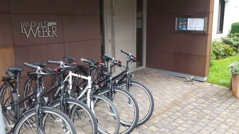 Een groep fietsen staat voor een ingang. De vloer is geplaveid en op de achtergrond is een bord zichtbaar.