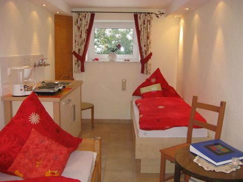 A cozy room with two single beds and red cushions. The room has a small kitchenette and a window with a view of the outdoors.