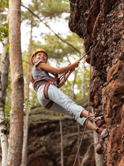 A happy child in a climbing helmet is climbing a rock wall. Surrounded by trees, they are enjoying the adventure in nature.