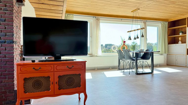 Een moderne woonkamer met een oranje sideboard en een grote televisie. De ruimte heeft grote ramen met uitzicht op het groen en een eettafel met stoelen.