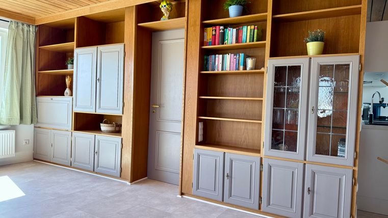 Een lichte ruimte met moderne, ingebouwde planken en een houten plafond. De planken zijn deels gesloten en deels open en herbergen boeken en decoratieve elementen.