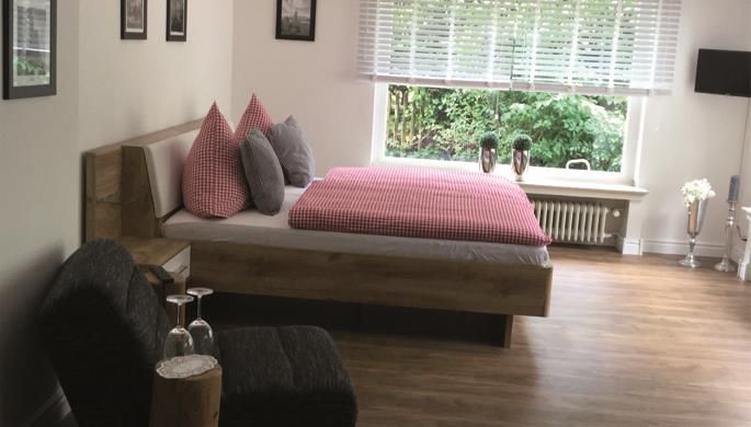 A modern bedroom with a cozy bed and checkered pillows. Large windows provide plenty of daylight and a view of the greenery.