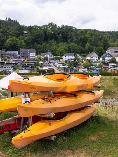 Three stackable orange kayaks are standing on a meadow. In the background, small houses and trees can be seen.