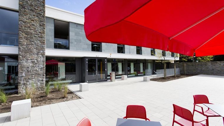 A modern courtyard with red umbrellas and tables. The adjacent facade of the building features large windows.
