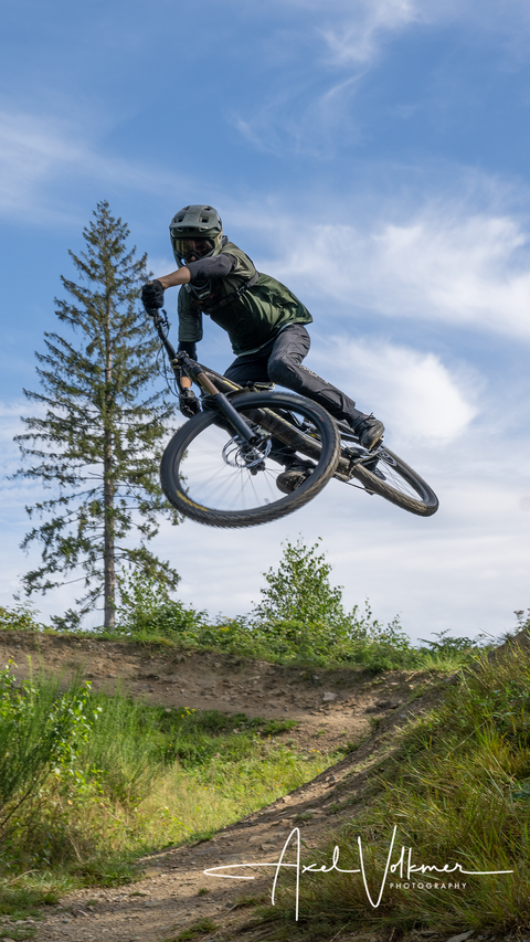 Een mountainbiker springt met zijn fiets over een heuvel. Op de achtergrond zijn bomen en een blauwe lucht te zien.